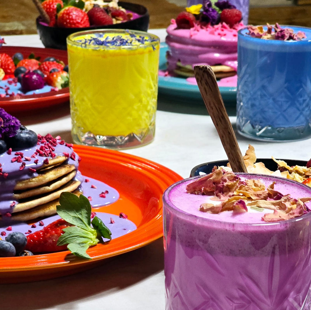 A tall glass filled with a vibrant blue spirulina latte, topped with edible flower petals and berries . Served on a table with colorful superfood drinks and pancakes in the background 