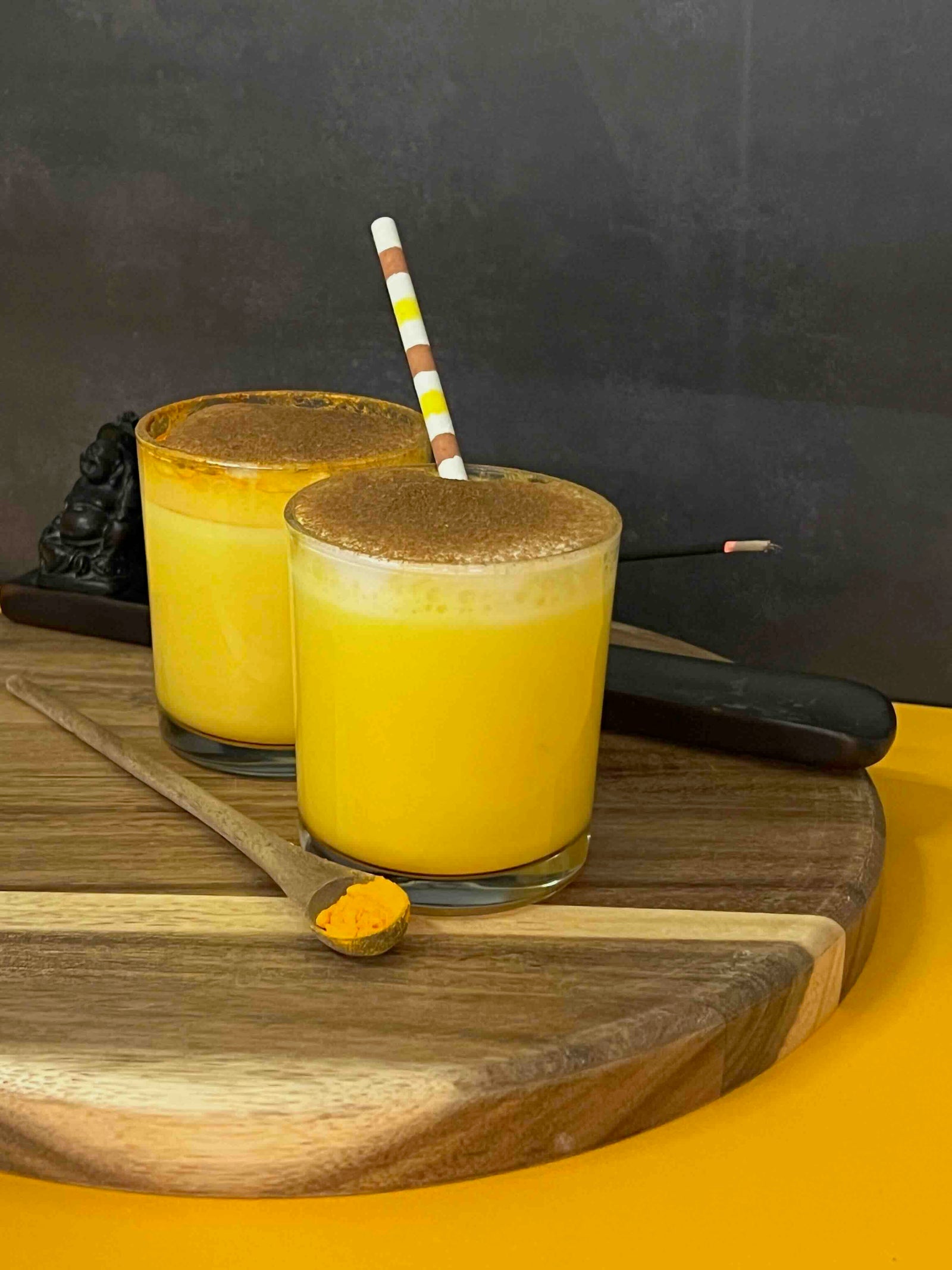 Two glasses of yellow smoothie with a brown topping on a wooden board, against a dark background.