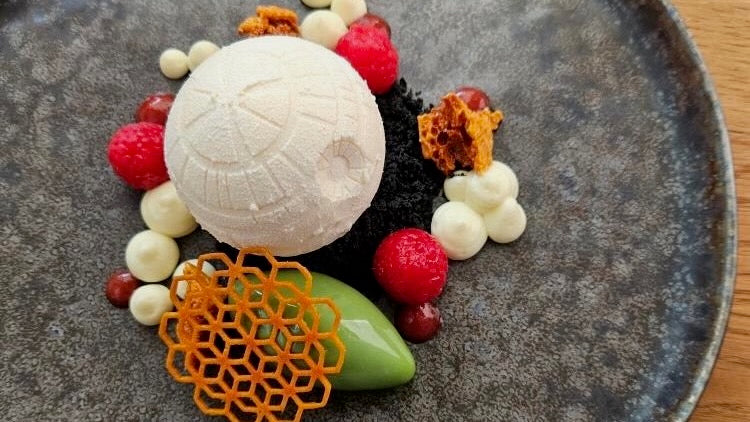 Delicate dessert with white sphere, berries, and decorative elements on a textured plate.