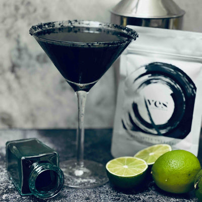 Black cocktail in a martini glass with lime wedges and a bottle on a dark surface.