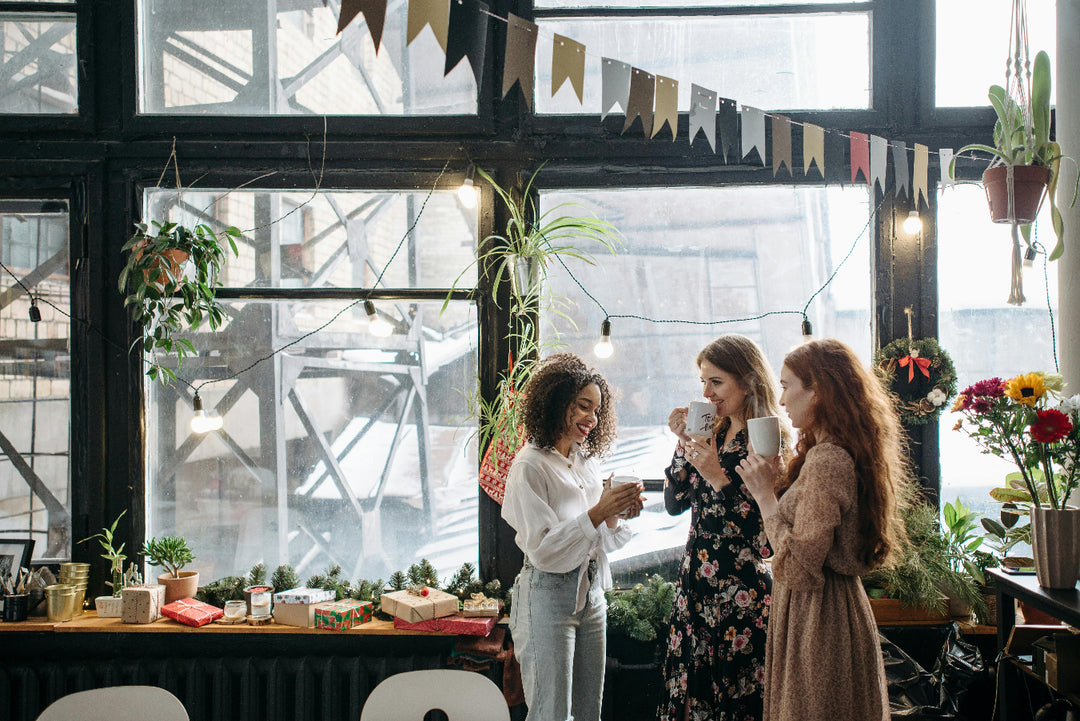 Three women standing in a room with large windows, plants, and a festive banner.