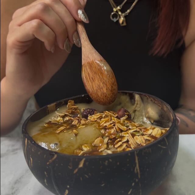 Hojicha smoothie bowl in a coconut bowl, topped with granola and dried fruit, with a wooden spoon held above it