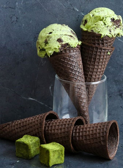 Green matcha ice cream in chocolate cones with a glass of ice cream in the background on a dark surface.