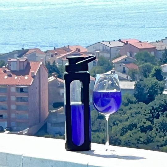 Yes Superfood glass shaker with black silicone sleeve beside a stemmed glass, both containing a vivid blue drink, with a sea view.
