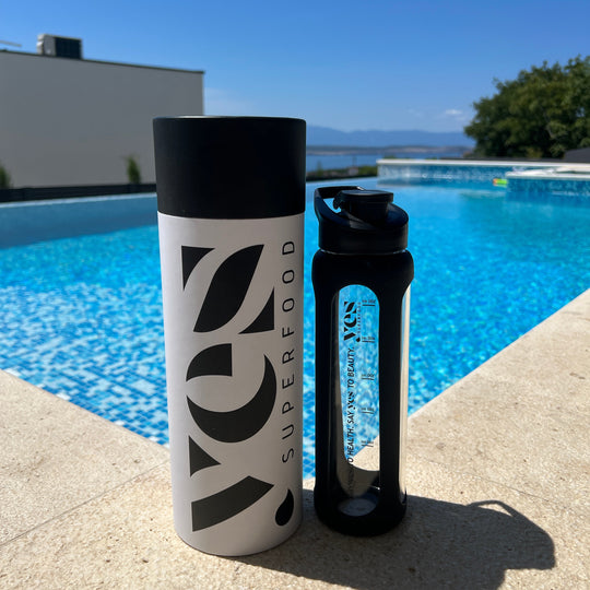 Yes Superfood glass shaker with black silicone sleeve beside its cylindrical gift box at the edge of a swimming pool.
